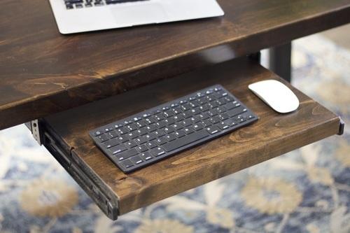The Standard Desk Add-On by James+James is displayed with a retractable keyboard tray. The tray accommodates a slim, black keyboard and a white wireless mouse. The desk also supports an open laptop, partially visible. The background includes a patterned rug.