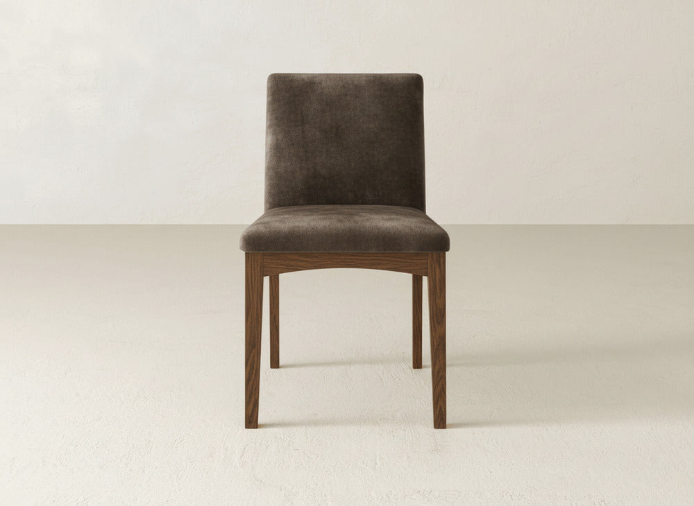 The James & James Klein Dining Chair has maple wooden legs and a brown upholstered seat, displayed on a light floor with an off-white wall backdrop.