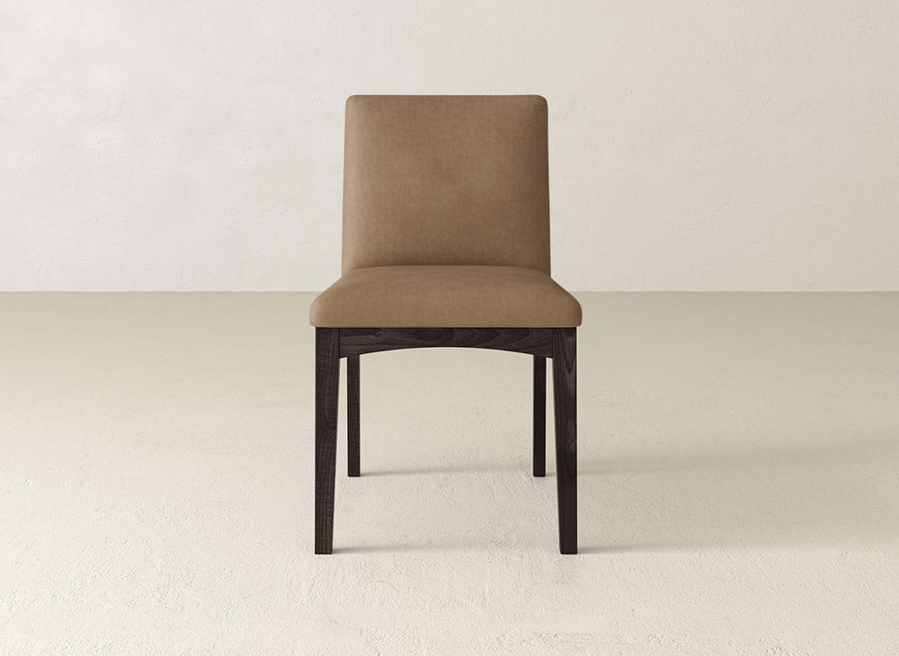 The James & James Klein Dining Chair has a brown upholstered seat and backrest with dark maple wooden legs, shown on a light floor against a white wall.
