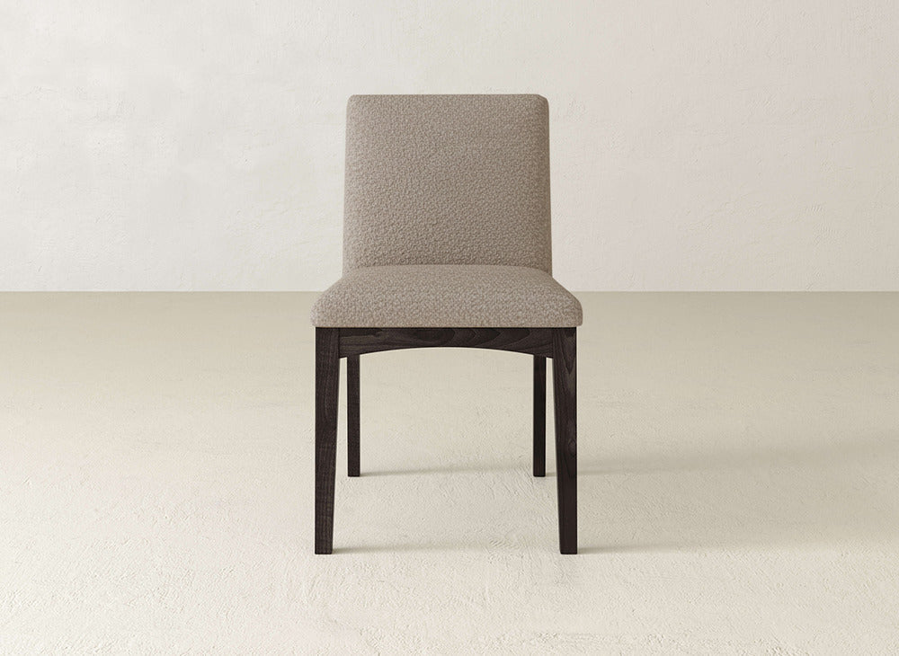 The James & James Klein Dining Chair features a light beige upholstered seat and backrest with elegant maple wood legs, shown against an off-white wall on a light-colored floor.