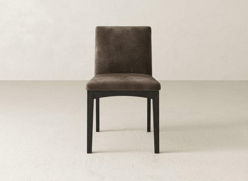 The Klein Dining Chair by James & James features a simple, modern design with a dark brown upholstered seat and backrest, complemented by four straight black wooden legs and shown against a light background.