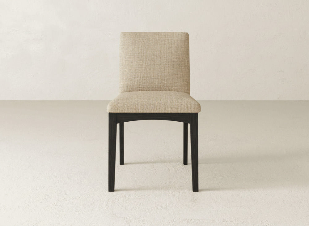 The James & James Klein Dining Chair has beige upholstery, a straight backrest, and sturdy maple legs. It’s shown on a light floor with a plain off-white wall behind.