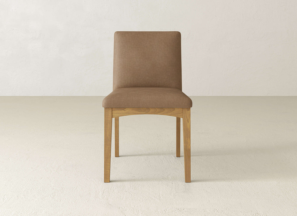 The James & James Klein Dining Chair, with a light brown cushioned seat and backrest and maple wooden legs, is placed on a light floor against an off-white wall.