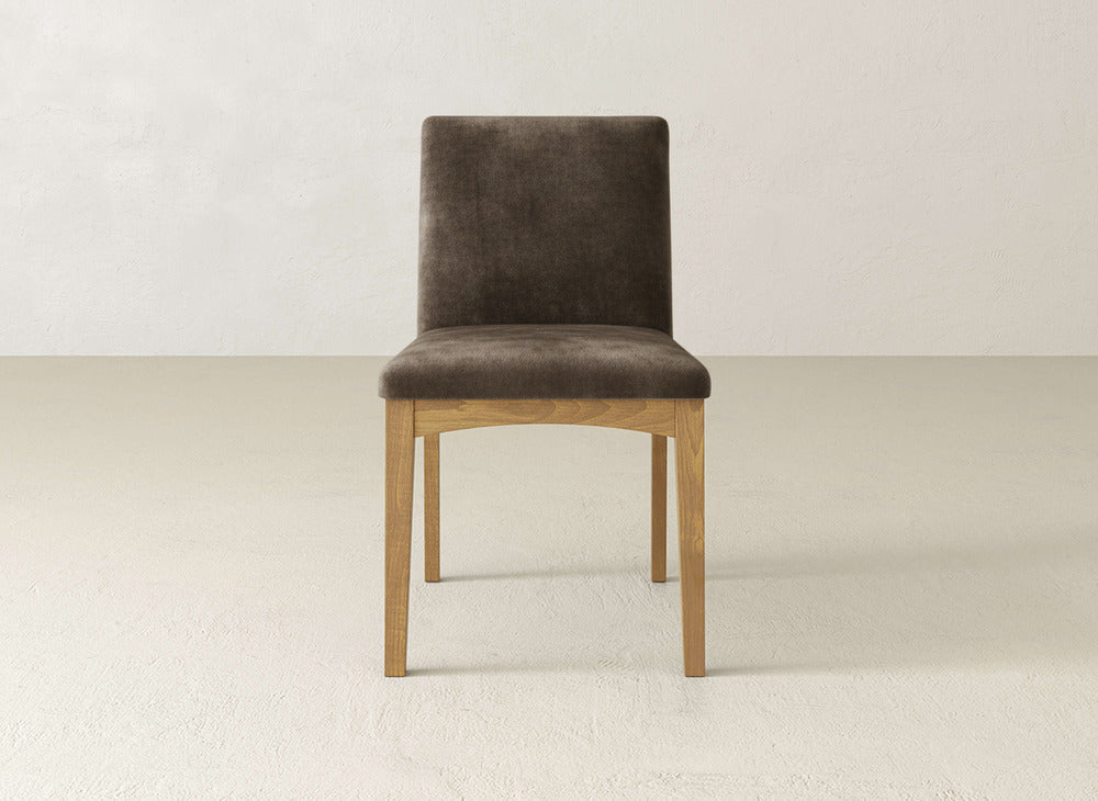 The Klein Dining Chair by James & James features brown upholstery and maple wooden legs, shown facing forward against a plain, light-colored wall and floor.