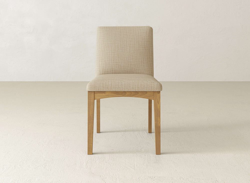 A James & James Klein Dining Chair with maple wooden legs and a light beige upholstered seat and backrest is centrally placed against a plain, off-white wall and floor.
