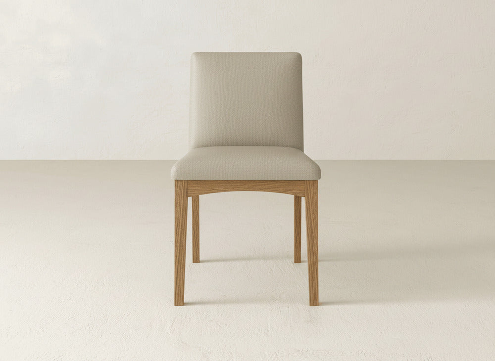 The James & James Klein Dining Chair features a simple beige design with a straight backrest and light maple wooden legs, shown on a light floor against an off-white wall.