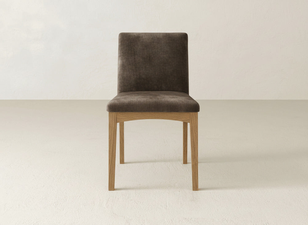 The Klein Dining Chair by James & James features brown upholstery and maple wooden legs, shown front-facing on a light floor against an off-white wall.