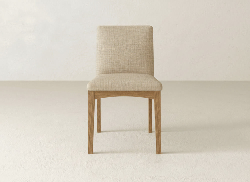 The Klein Dining Chair by James & James, featuring beige upholstery and maple wooden legs, is centered in a minimalist room with light-colored walls and flooring.