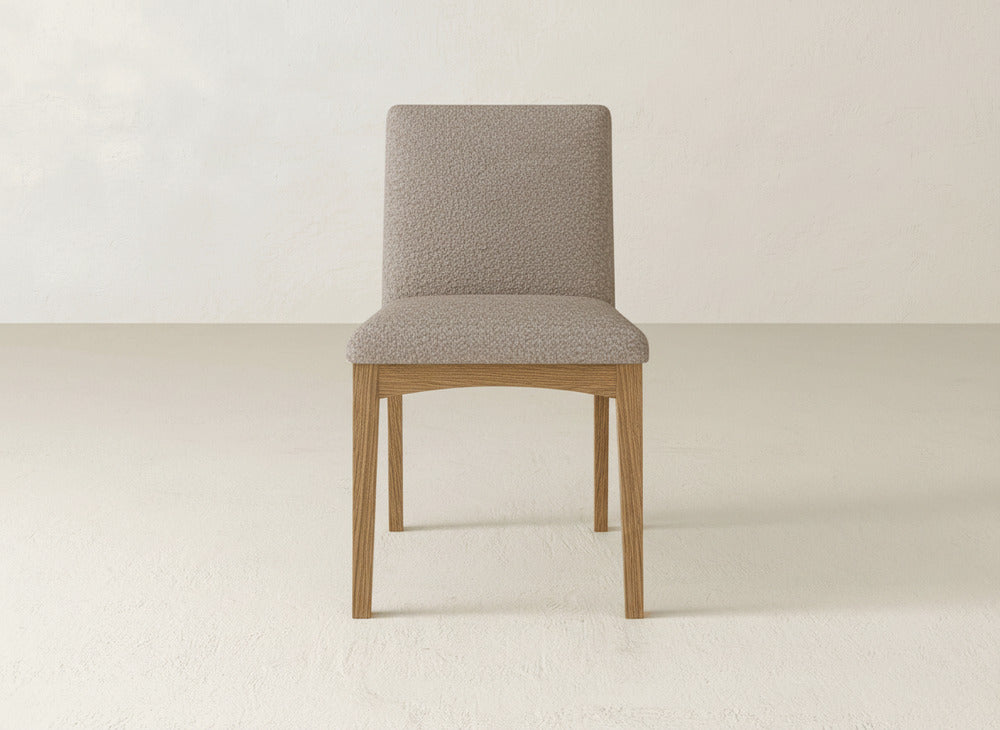 The James & James Klein Dining Chair features a light beige upholstered seat and backrest with elegant maple wooden legs, shown against a plain off-white background.