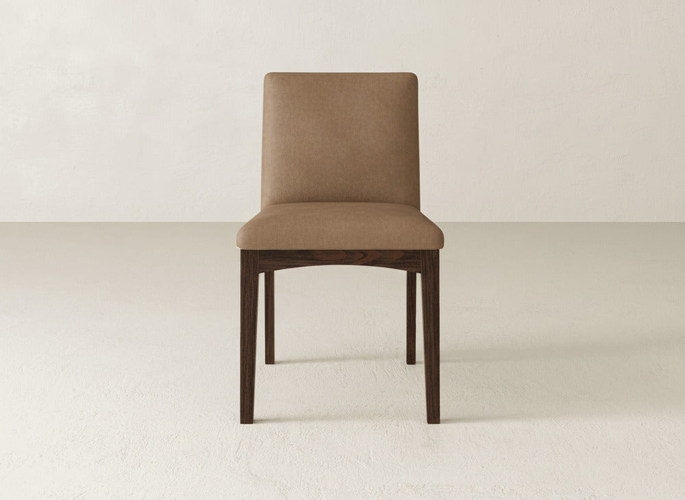 The James & James Klein Dining Chair features a padded light brown seat and backrest with dark maple legs, set against a plain white wall and floor.