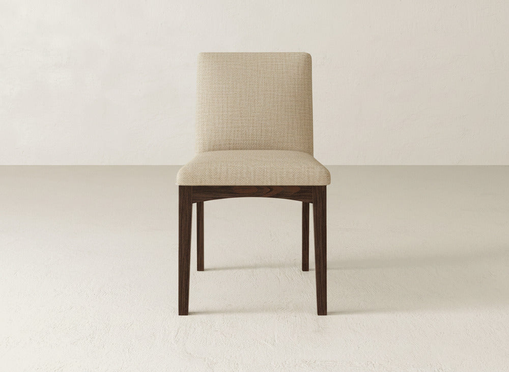 A James & James Klein Dining Chair with a beige upholstered seat and backrest and dark maple wooden legs sits in the center of a plain, light-colored room.