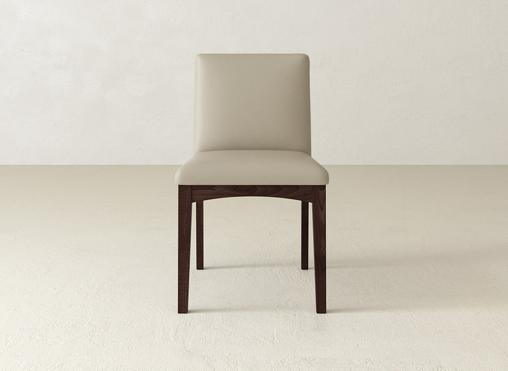 The Klein Dining Chair by James & James features a light beige upholstered seat and backrest with dark maple legs, set on a light floor against an off-white wall.