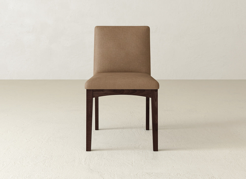 The James & James Klein Dining Chair features a light brown upholstered seat and backrest with dark maple legs, set against an off-white textured wall and floor.