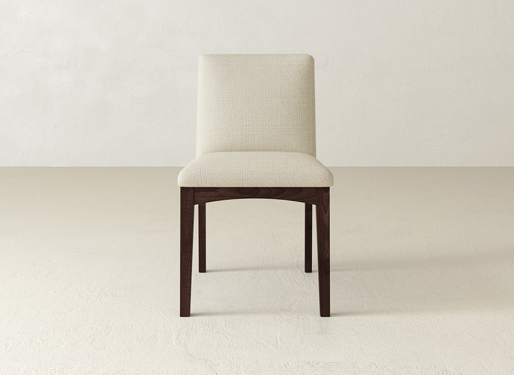 The Klein Dining Chair by James & James features a cushioned, cream-upholstered seat and backrest with dark maple legs, shown on a light floor against an off-white wall.