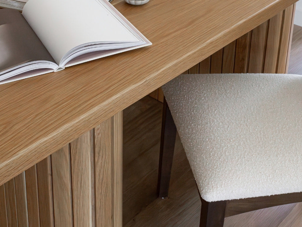 A close-up of a wooden desk with an open book and a James+James Bisque - Boucle Swatch chair tucked underneath, both exuding casual elegance on a wooden floor.