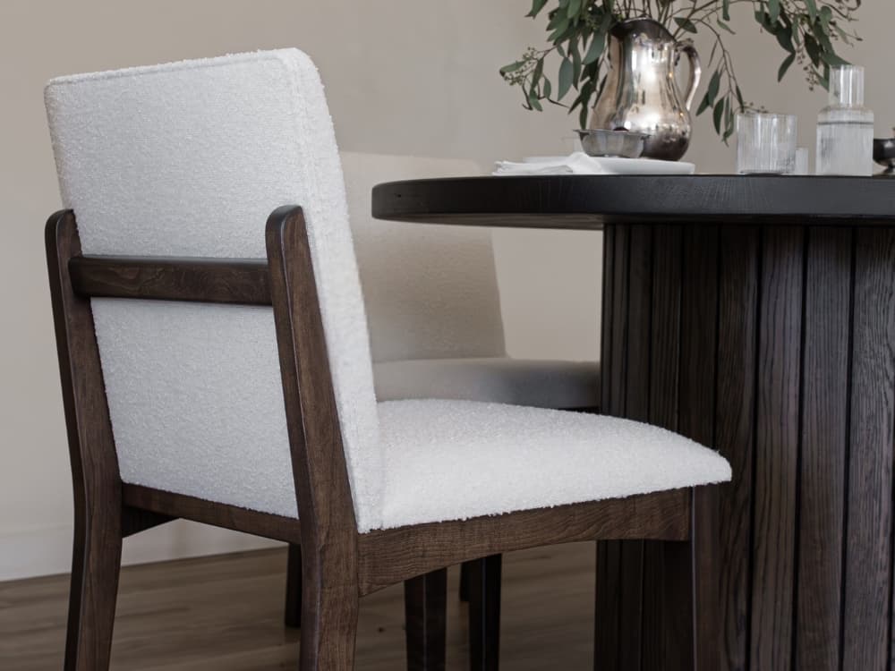 A close-up of two modern chairs featuring James+James Bisque - Boucle upholstery and dark wooden frames, placed by a round dark wood table set with a silver pitcher and glassware, exuding casual elegance.