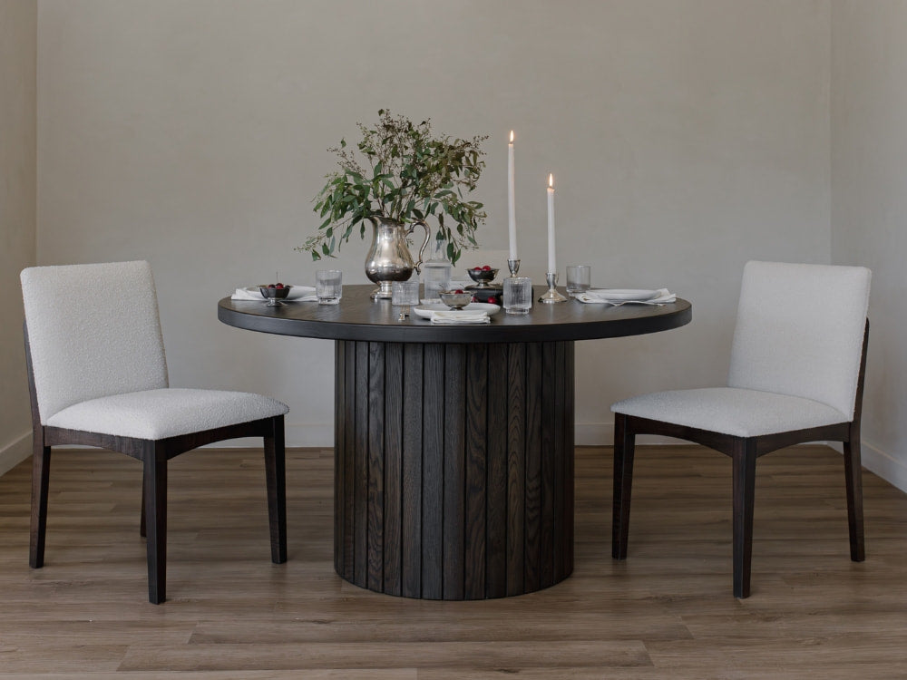 A round dark wood dining table set for two with James+James Bisque - Boucle chairs, white plates, glasses, and lit candles. A vase with greenery centers the minimalist table, adding casual elegance to the space.