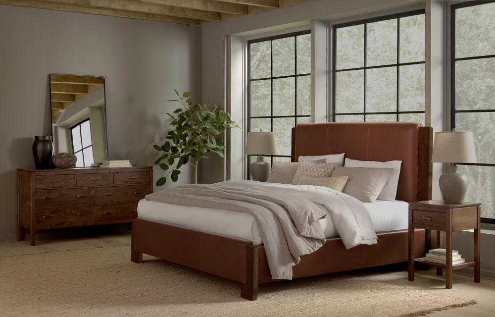 In a modern bedroom, the James & James Lennon Bed in brown linen features an upholstered headboard and white/gray bedding, flanked by maple nightstands with lamps. A large dresser with a mirror and vases sits to the left, while a leafy plant and big windows complete the cozy space.