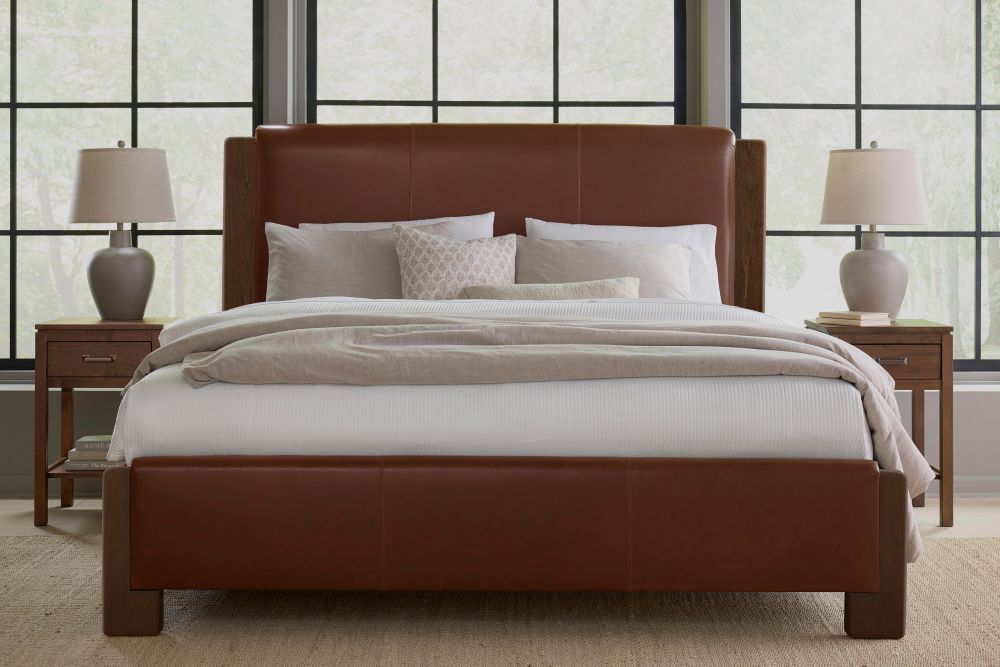 The bedroom showcases the Lennon Bed – Linen by James & James, featuring an upholstered headboard and footboard. Beige and white bedding with pillows add to the design. Nightstands with lamps flank the bed, while large windows offer views of tall trees and maple wooden accents provide warmth.