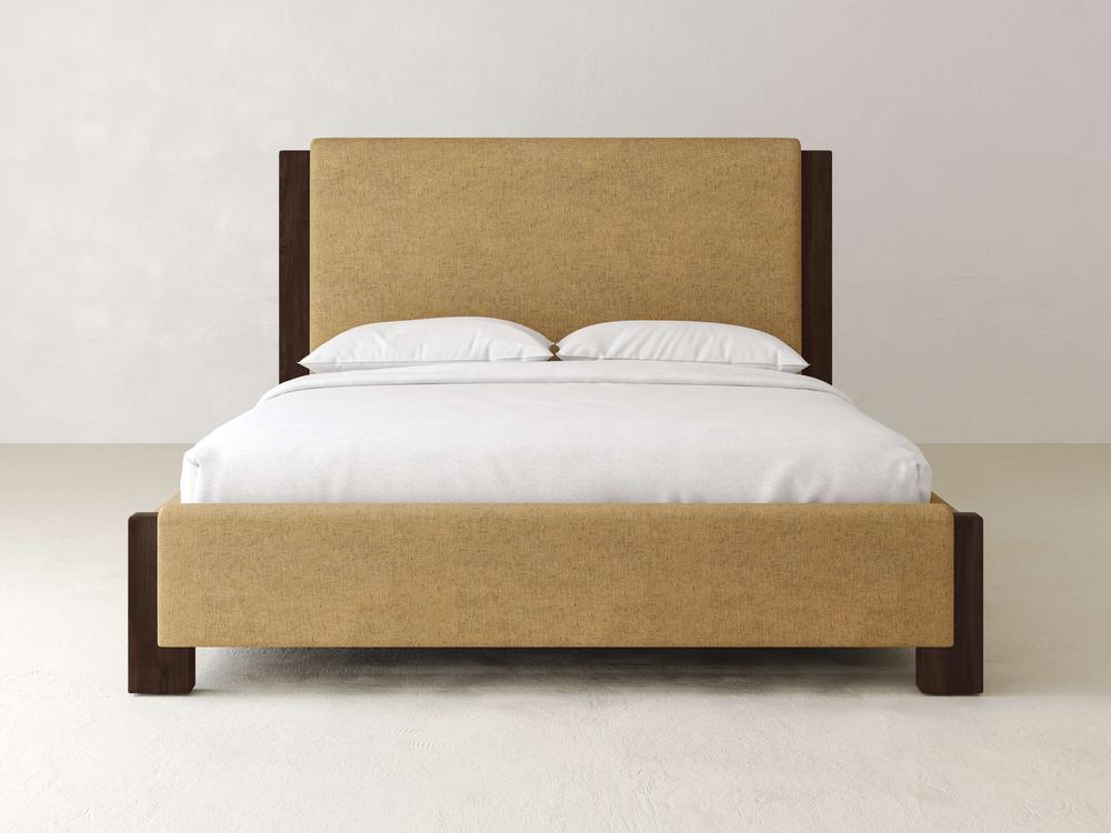 The Lennon Bed – Linen by James & James is a modern piece with a tall upholstered headboard made from performance fabric, featuring maple wooden accents on its frame. It includes a white sheet and two pillows, and is set against a plain light-colored wall and floor.