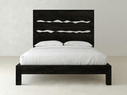 The James+James Live Edge Bed in Charred Ember on Alder features a modern dark wood frame with a high headboard and two wavy horizontal cutouts, topped with white bedding and two pillows, set against a light-colored wall.