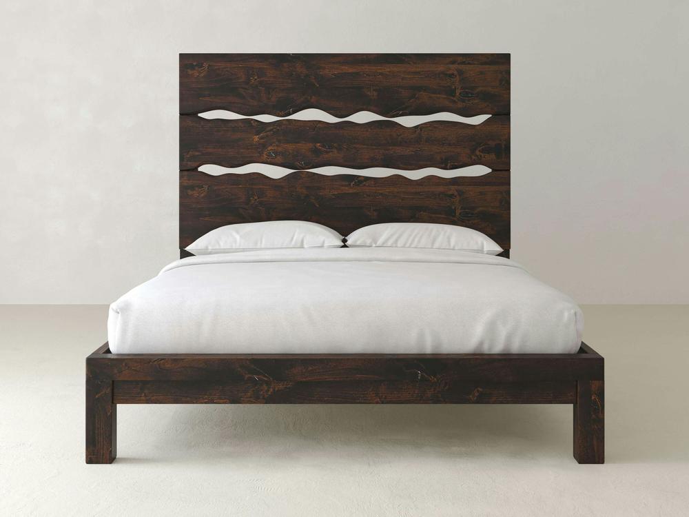 The James+James Live Edge Bed in Tobacco on Alder features a tall, wavy-patterned headboard and is shown with simple white bedding against a plain, light-colored wall.