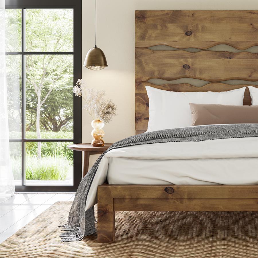 A cozy bedroom with a wooden bed frame and headboard, dressed in white and beige linens. A gray throw is draped over the edge. A wooden nightstand with a lamp and decorative items sits beside a large window with a view of greenery outside.