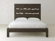 The James+James Live Edge Bed – Espresso on Oak features a tall, dark brown headboard with three wavy cut-out designs, dressed in white bedding and pillows, set against a plain light-colored wall and floor.