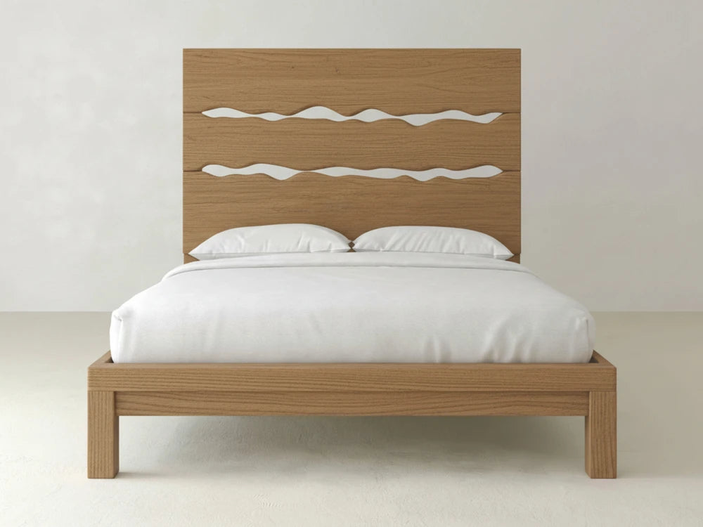 The James+James Live Edge Bed - Oat on Oak is a wooden bed frame with a tall headboard featuring three wavy cut-out designs, styled with white pillows and bedspread against a plain white wall.
