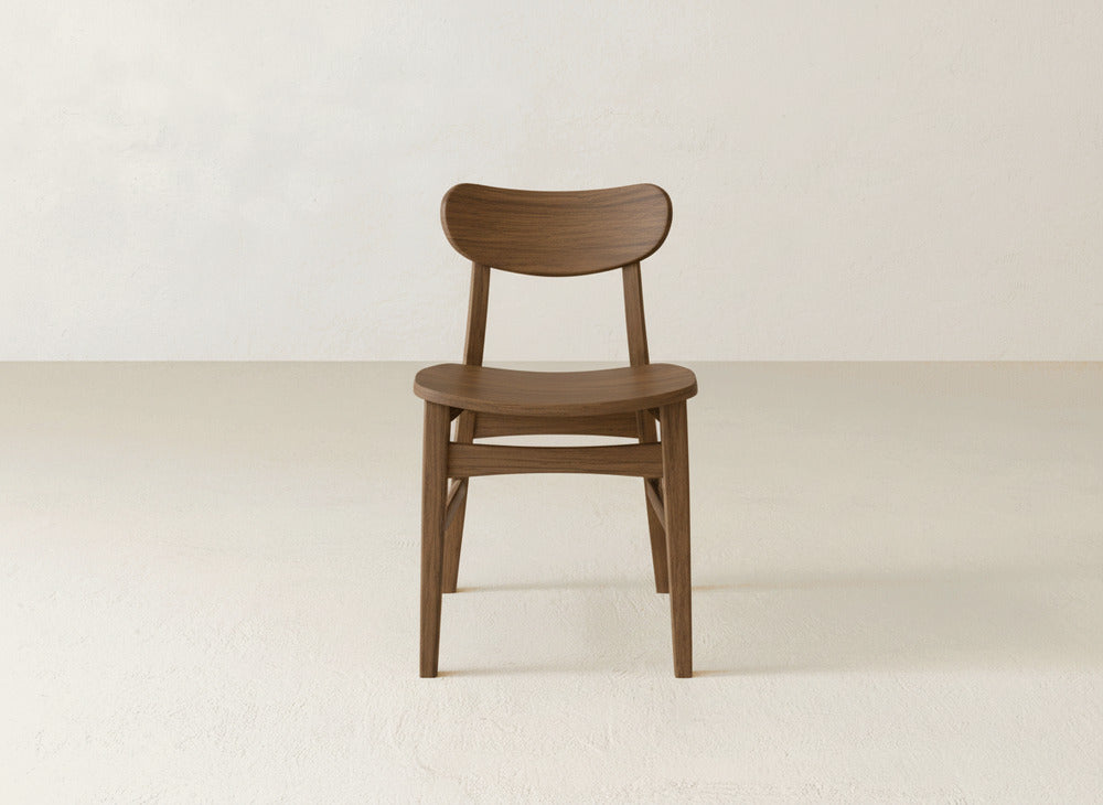 The Lynn Dining Chair by James+James is crafted from solid maple hardwood with a curved backrest and seat, displayed on a light floor and an off-white wall—a timeless piece of American craftsmanship.