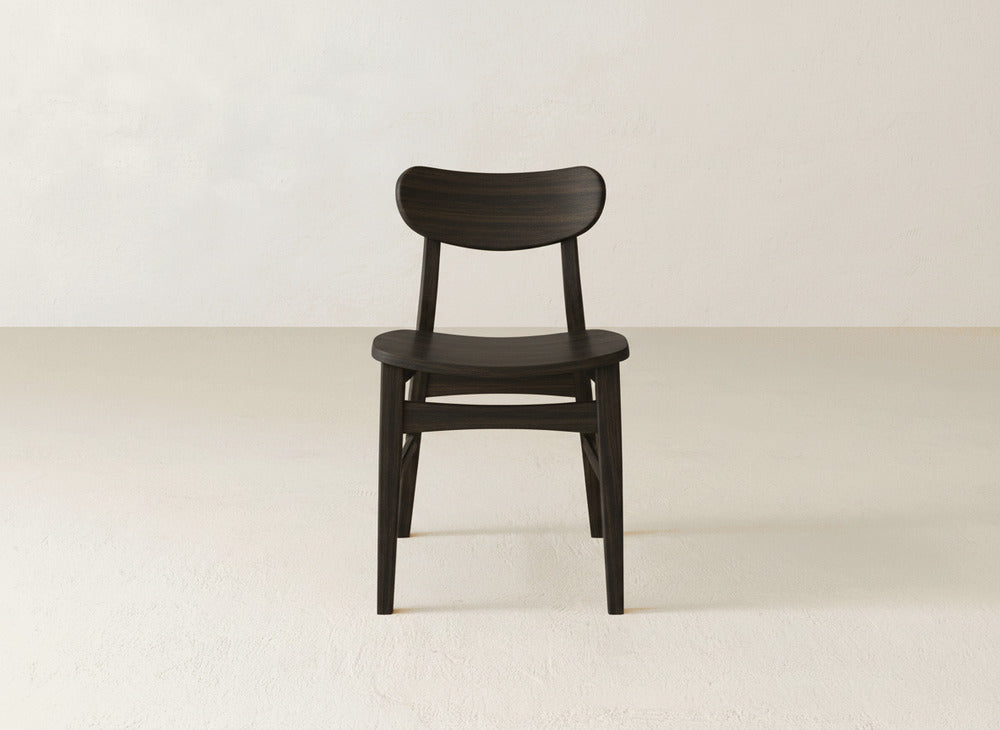 A James+James Lynn Dining Chair, featuring a dark solid maple build, curved backrest, and minimalist design, sits on a light floor against an off-white wall.