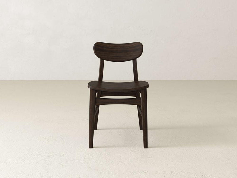 The Lynn Dining Chair from James+James, crafted from maple wood, showcases a minimalist design with a curved backrest and geometric legs. It features a dark finish and is set against an off-white textured wall in a simple, light-colored room.