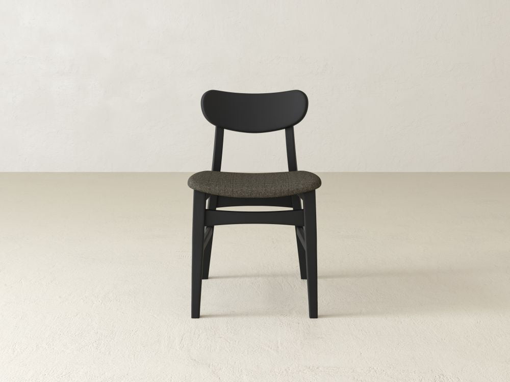 The James+James Lynn Upholstered Dining Chair in modern black with plush upholstery and a curved backrest sits elegantly on a neutral floor against a light wall, perfectly embodying the collection's minimalist aesthetic.