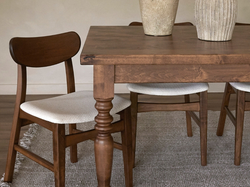 A wooden dining table with turned legs by James+James is surrounded by wooden chairs featuring Oatmeal - Textured Linen upholstered seats, arranged on a textured gray rug, with two decorative vases on the tabletop.