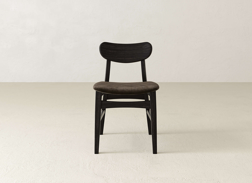 The Lynn Upholstered Dining Chair by James+James features a maple frame, curved dark backrest, and cushioned brown seat, showcasing modern sophistication on a light floor against an off-white wall.