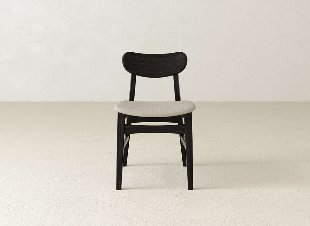 The James+James Lynn Upholstered Dining Chair features a curved dark wooden backrest and light upholstered seat, exuding modern sophistication on a light floor against an off-white wall.
