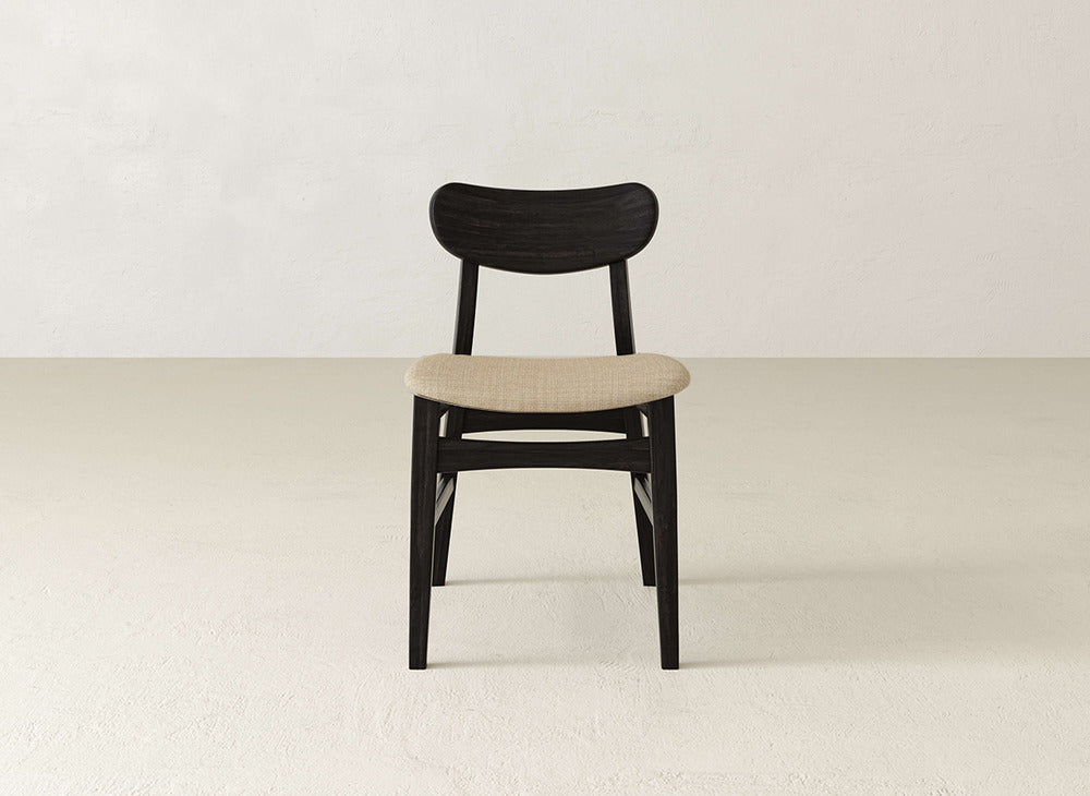 The Lynn Upholstered Dining Chair by James+James features a modern curved dark brown backrest, light beige fabric seat, and sleek maple wood accents, shown on a light floor against an off-white wall.