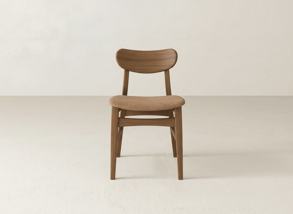 The Lynn Upholstered Dining Chair by James+James features a curved backrest and cushioned seat in maple wood, set on a light floor against a plain off-white wall.