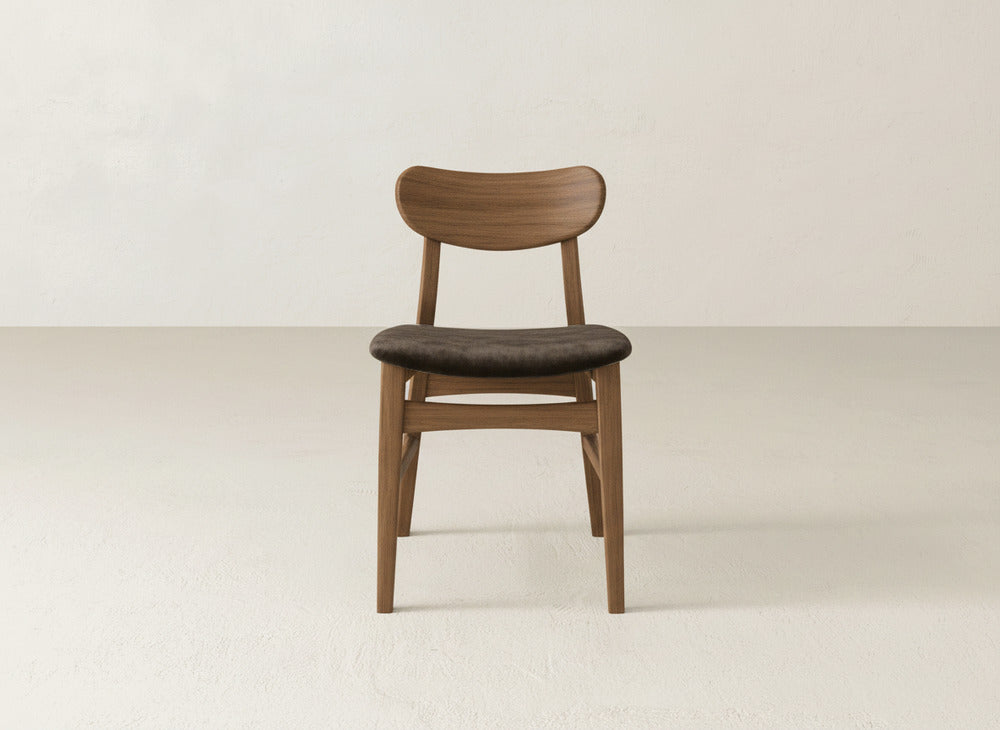 The Lynn Upholstered Dining Chair by James+James, featuring a curved maple backrest and dark cushioned seat, brings modern sophistication to minimalist spaces with light floors and walls.