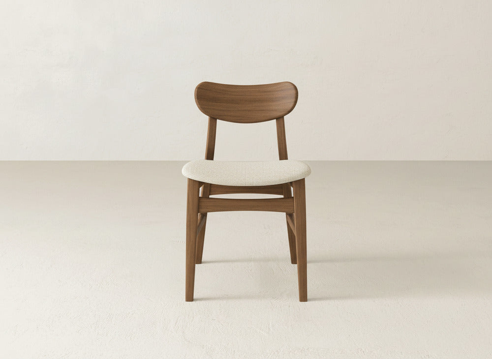 The Lynn Upholstered Dining Chair by James+James, featuring a curved backrest and light seat, stands on a pale floor beside an off-white wall, showcasing modern sophistication with its sleek, elegant maple wood design.