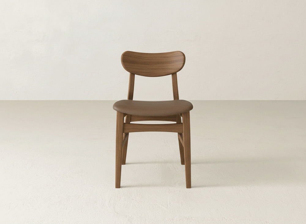 The Lynn Upholstered Dining Chair by James+James features a minimalist, modern design with a curved backrest, showcased on a light floor against an off-white wall for sophisticated appeal.