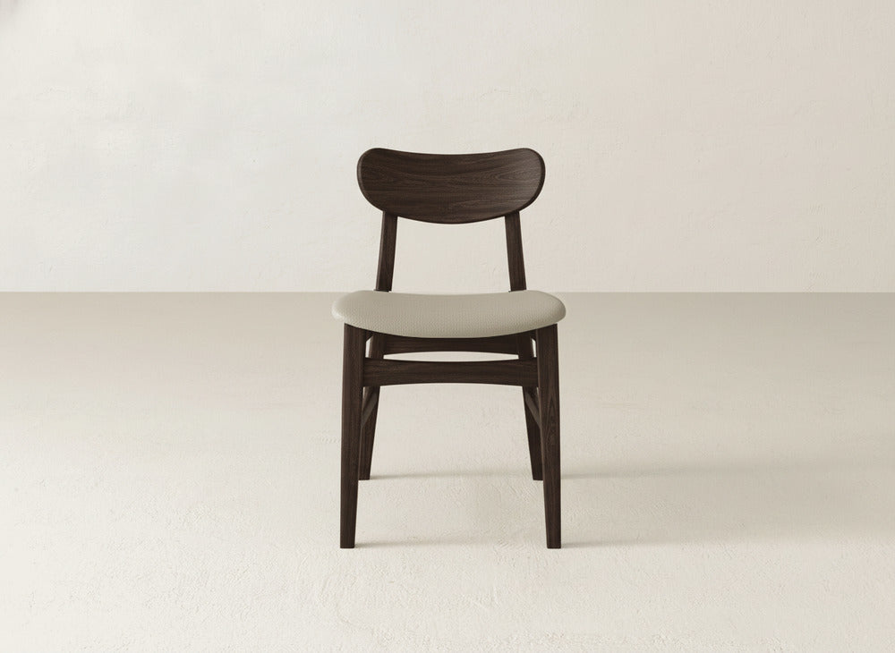 The Lynn Upholstered Dining Chair by James+James features a dark brown maple wood frame and a light beige cushioned seat, shown on a light floor with an off-white wall backdrop.