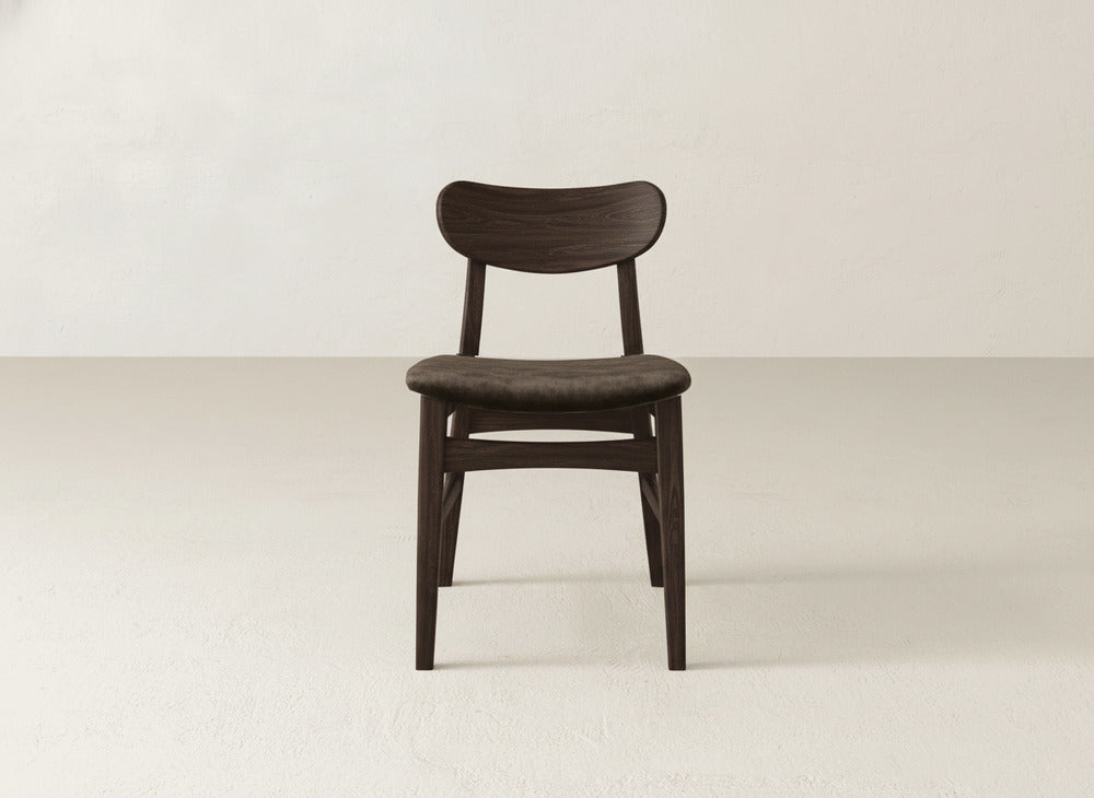 The Lynn Upholstered Dining Chair by James+James features a cushioned seat and curved backrest in dark maple, adding modern sophistication to any light-colored room with an off-white wall.