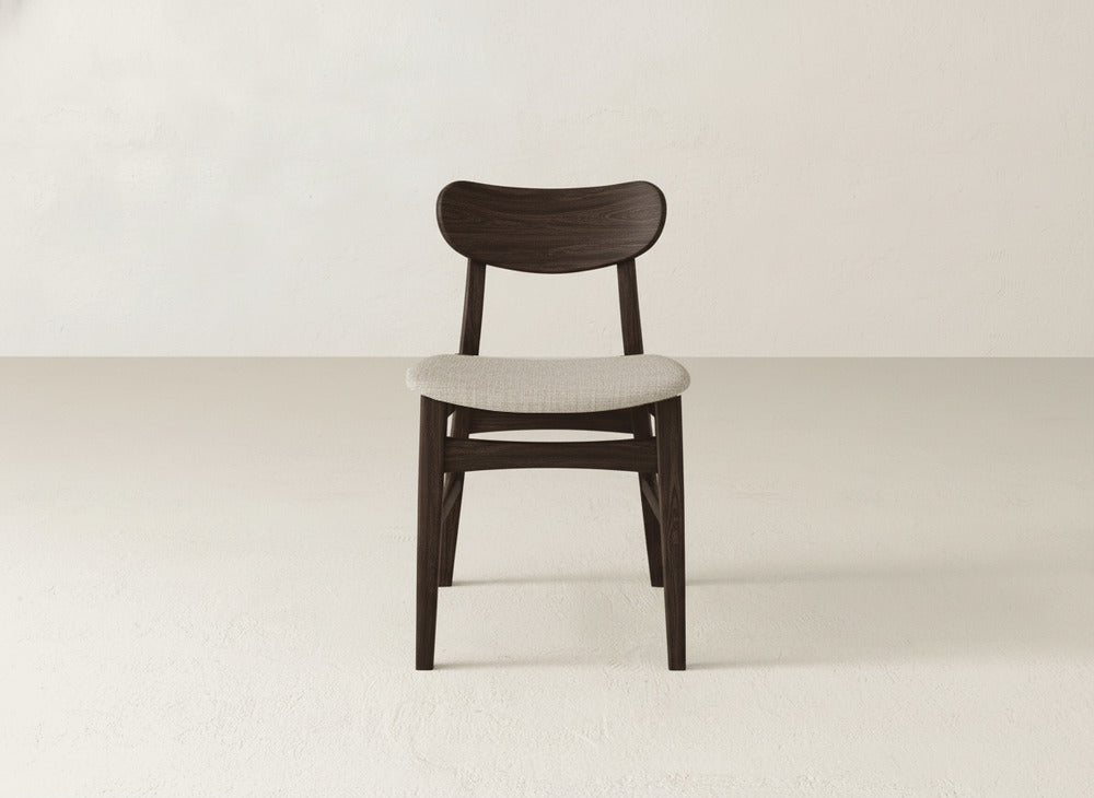 The Lynn Upholstered Dining Chair by James+James features a dark brown maple frame and light beige seat, showcasing modern elegance. It’s placed on a light floor against an off-white wall for a refined, sophisticated look.
