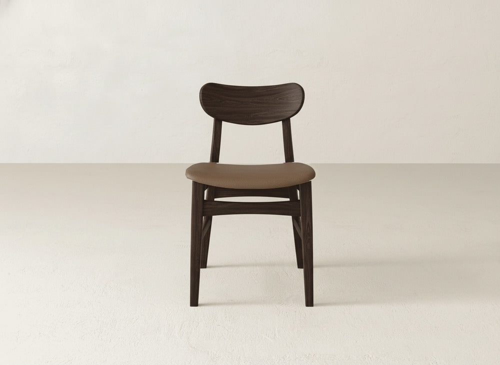The Lynn Upholstered Dining Chair by James+James, featuring a curved backrest and brown cushioned seat, stands out in a minimal, light-toned room with its dark maple wood frame contrasting the simple walls and floor.