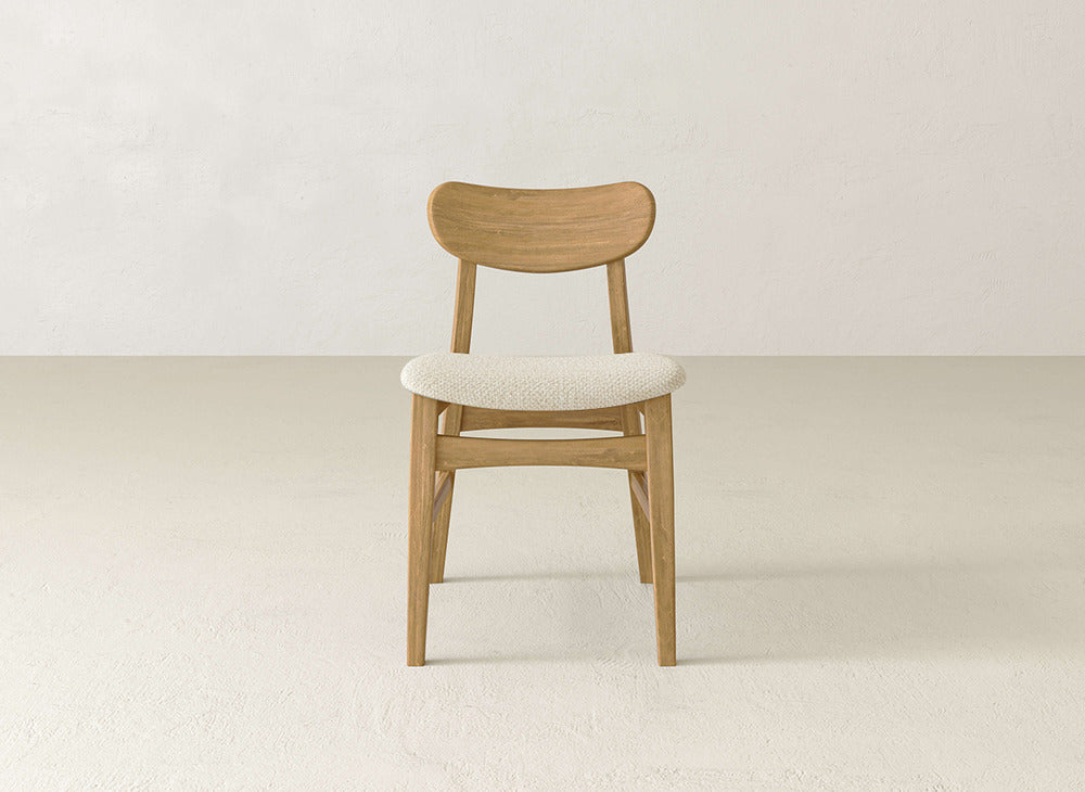 A James+James Lynn Upholstered Dining Chair, featuring maple wood, a curved backrest, and a light cushioned seat, is centered in an empty room with neutral walls and floor.