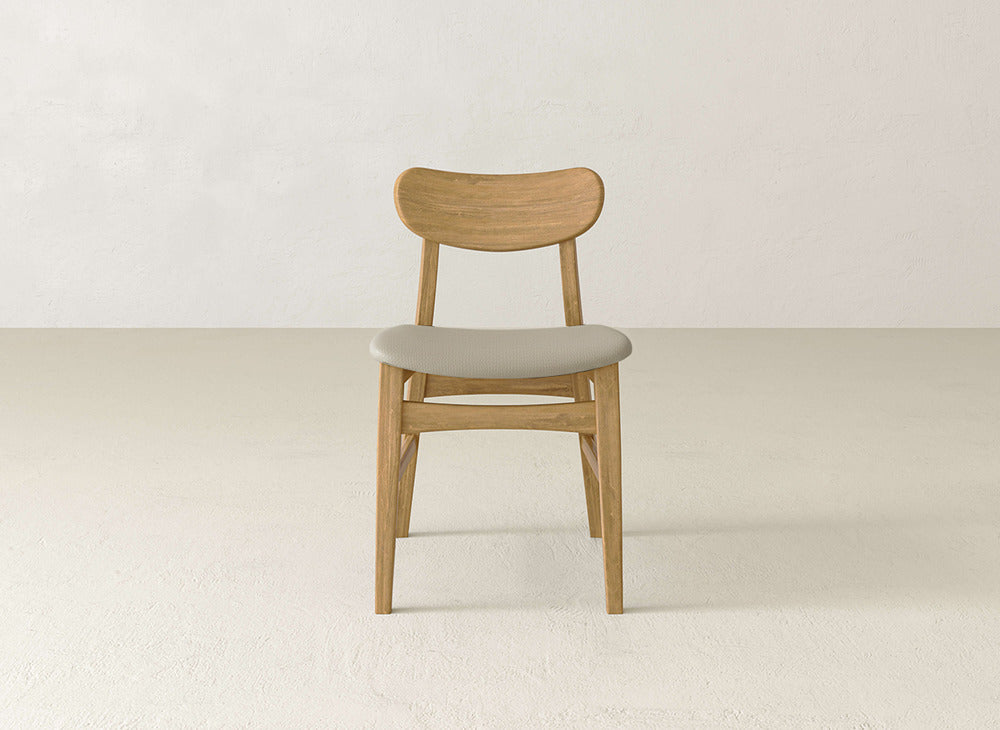 The James+James Lynn Upholstered Dining Chair, featuring maple wood, a curved backrest, and a light gray cushioned seat, is placed on a light floor against an off-white wall.