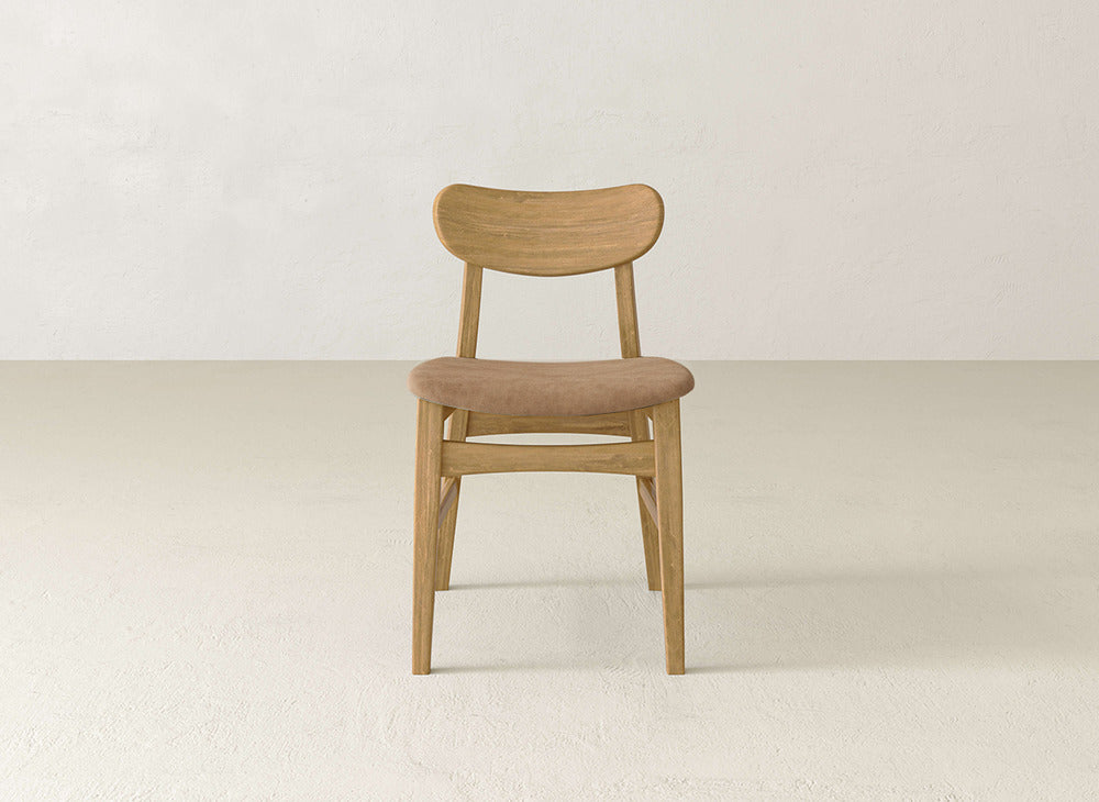 The Lynn Upholstered Dining Chair by James+James features a curved backrest and a light brown cushioned seat, crafted from maple wood, and is shown on a light floor with an off-white wall backdrop.