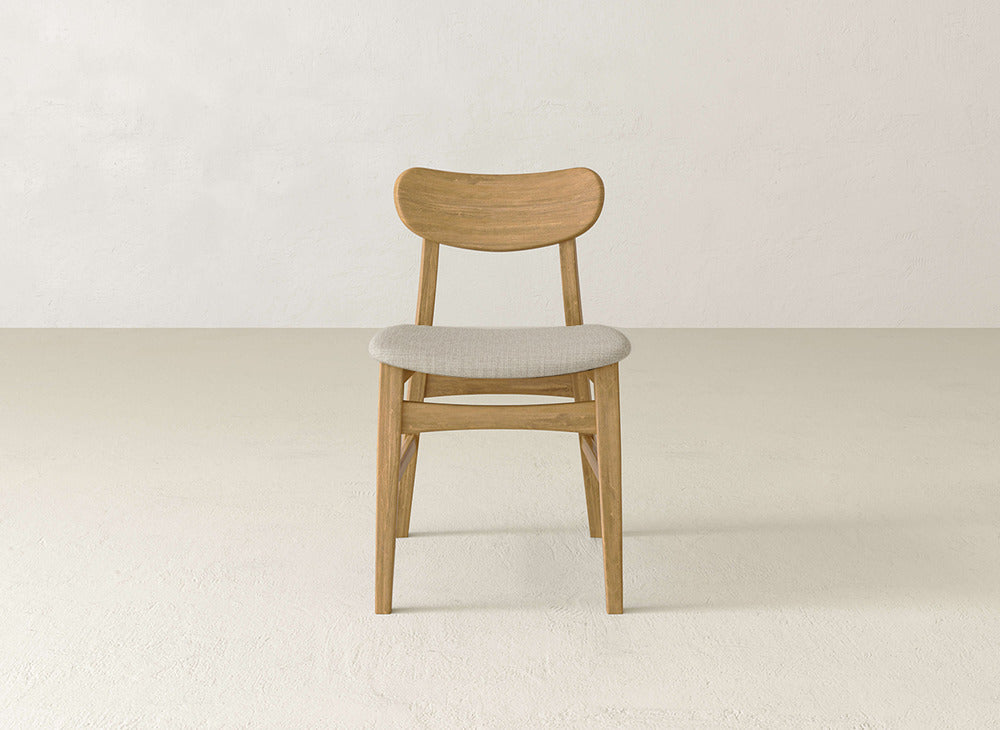 The James+James Lynn Upholstered Dining Chair has a curved backrest and light gray cushioned seat crafted from maple wood, set against a neutral textured wall—ideal as a modern dining chair.