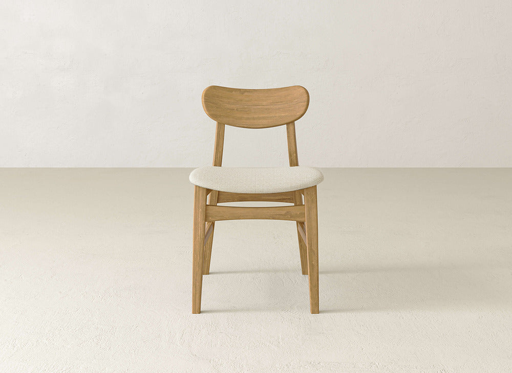 A James+James Lynn Upholstered Dining Chair with a curved backrest and light-colored cushion sits centered in a minimalist, empty room with light walls and flooring.